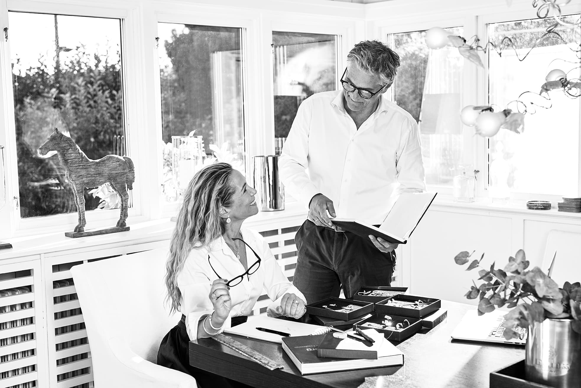 En kvinne sitter ved et skrivebord med smykker og holder et par briller. En mann står ved siden av henne med en åpen mappe. Begge befinner seg i et lyst og moderne rom.