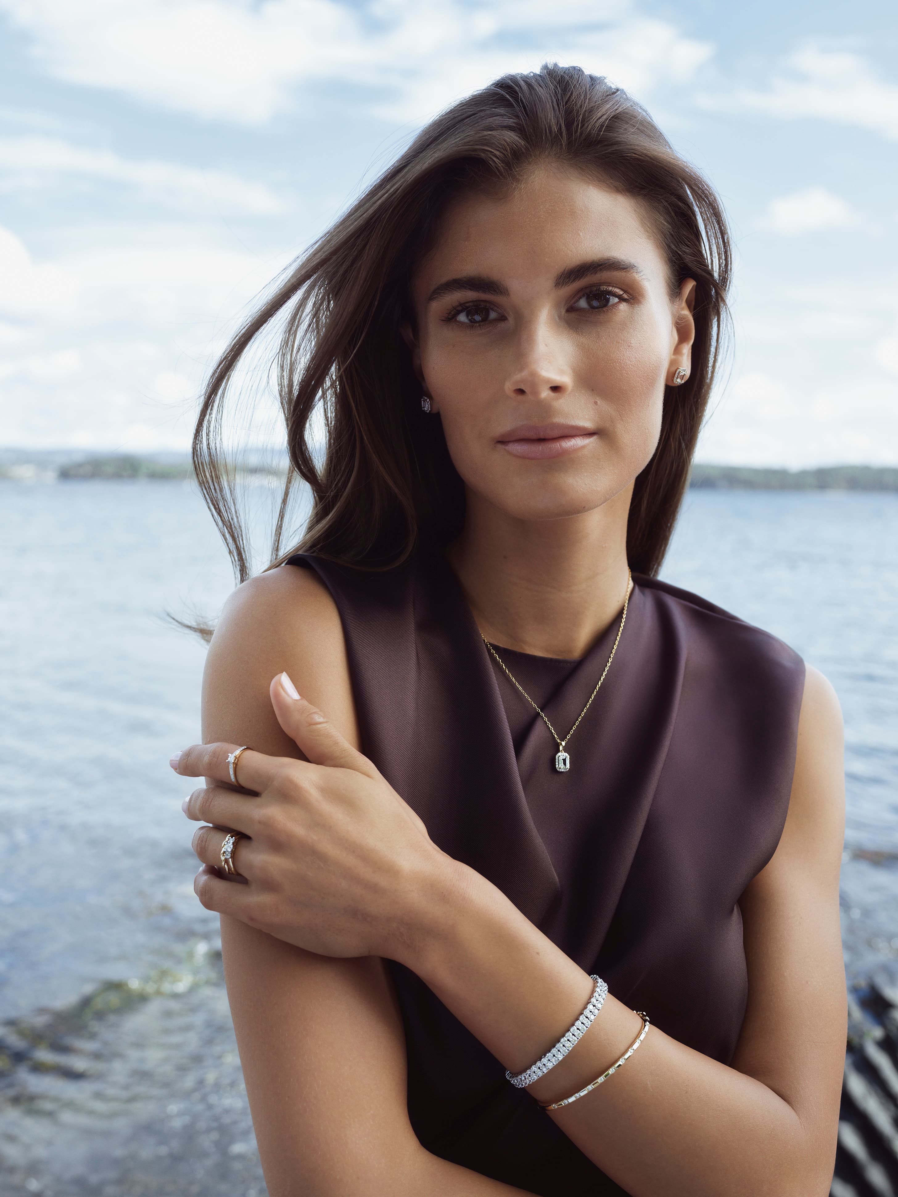 Kvinne ved en innsjø med vind i håret, iført en ermeløs topp og smykker, med et rolig uttrykk. Lys og skyet himmel i bakgrunnen.