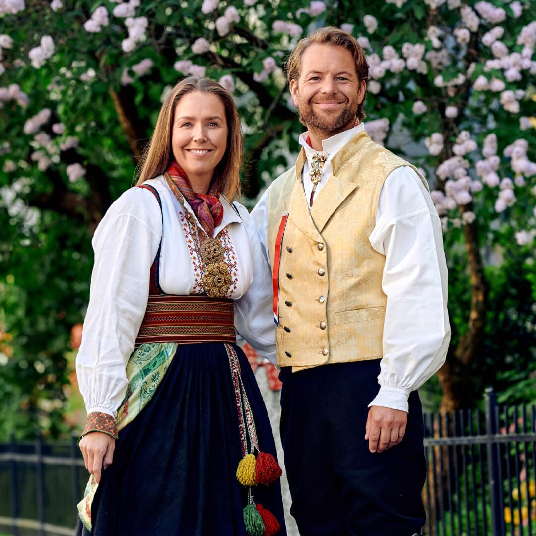 En mann og kvinne i tradisjonelle norske bunader, står smilende foran et blomstrende tre.