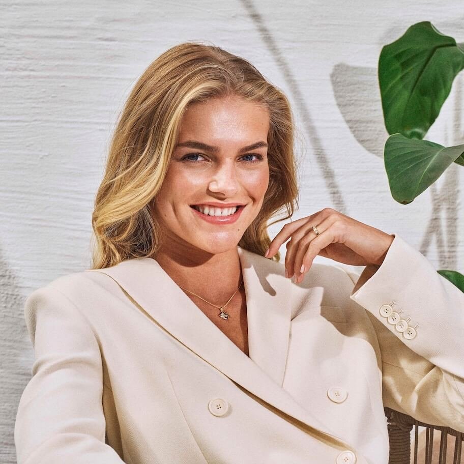 Les om gaver til henne Smilende kvinne med blondt hår og kremfarget dress viser frem gullsmykke og ring.