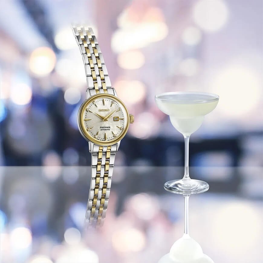 En tofarget Seiko-klokke som henger i løse luften ved et Martini-glass som står på et glassbord med lyse lillafarger og lys i bakgrunnen.