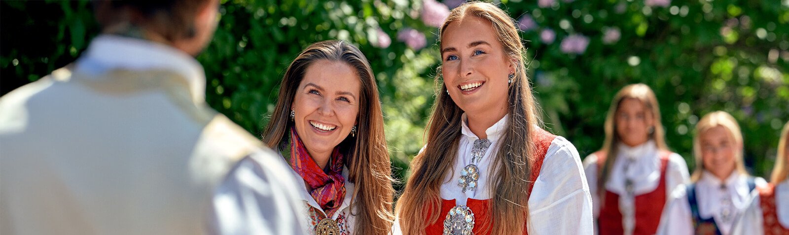 To kvinner i bunad smiler mot en mann i bunad, med to andre bunadkledde kvinner i bakgrunnen utendørs.
