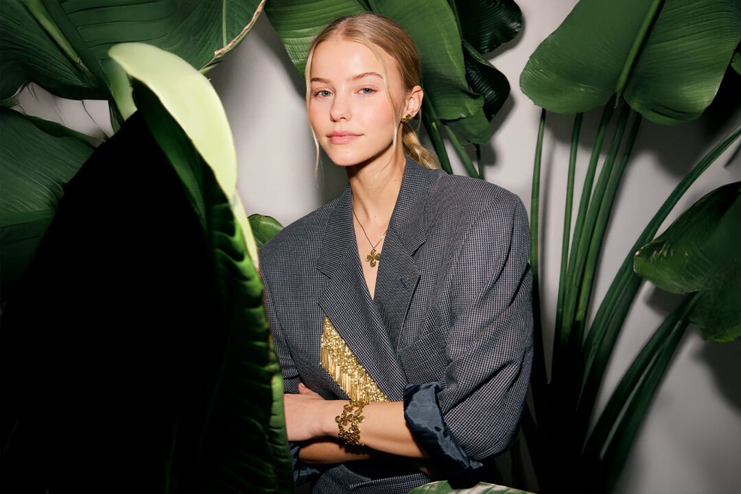 Halskjede – i gull eller sølv, med perler, symboler og plater Ung kvinne med lyst hår og grå blazer står blant store grønne blader, iført gullsmykker med detaljer og strukturert design.