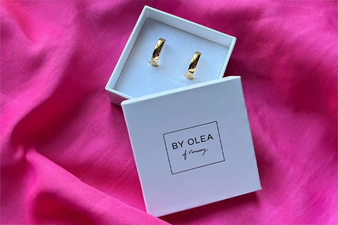 Se HEDDA øreringer Et par brede og gullbelagte stål øreringer HEDDA i en hvit gaveeske med By Olea of Norway på lokket, ligger oppå en rosa duk.