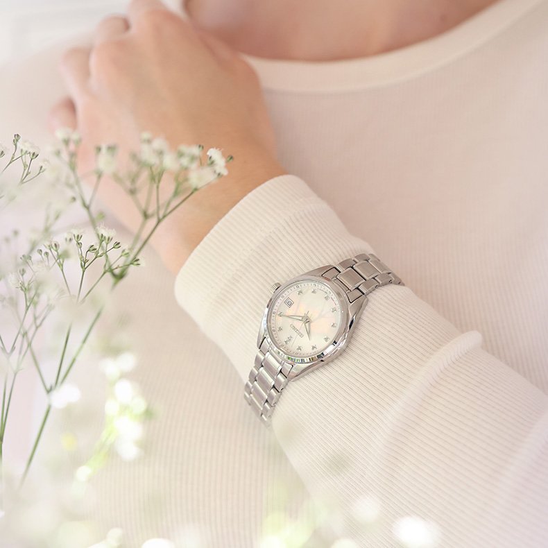 Les om dameklokker! Sølvfarget dameklokke med lenke og perlemorskive på håndledd, omgitt av myke blomster i lys setting.