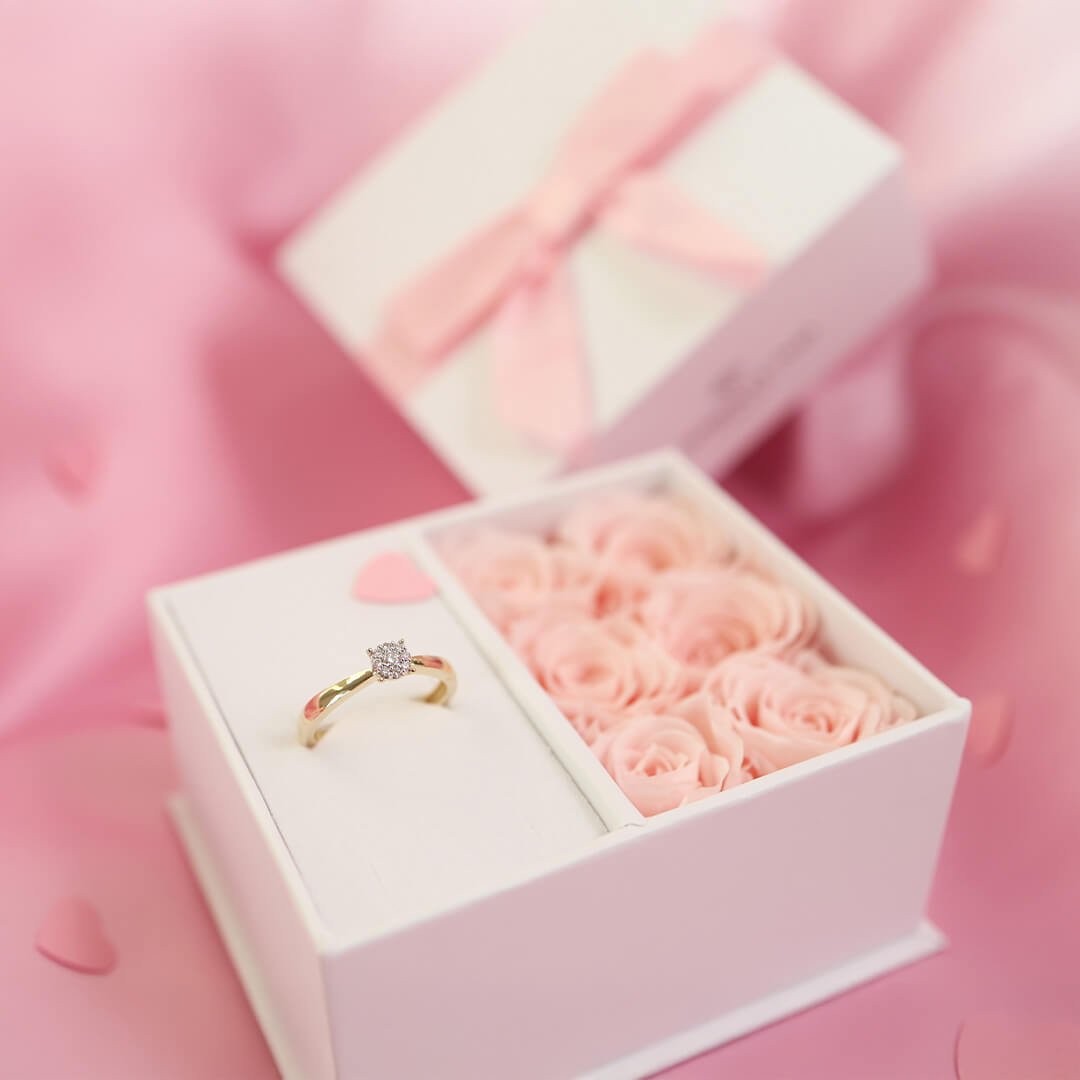Les om diamanter Gullring med diamant ligger i hvit gaveeske med rosa roser og sløyfe mot rosa bakgrunn.