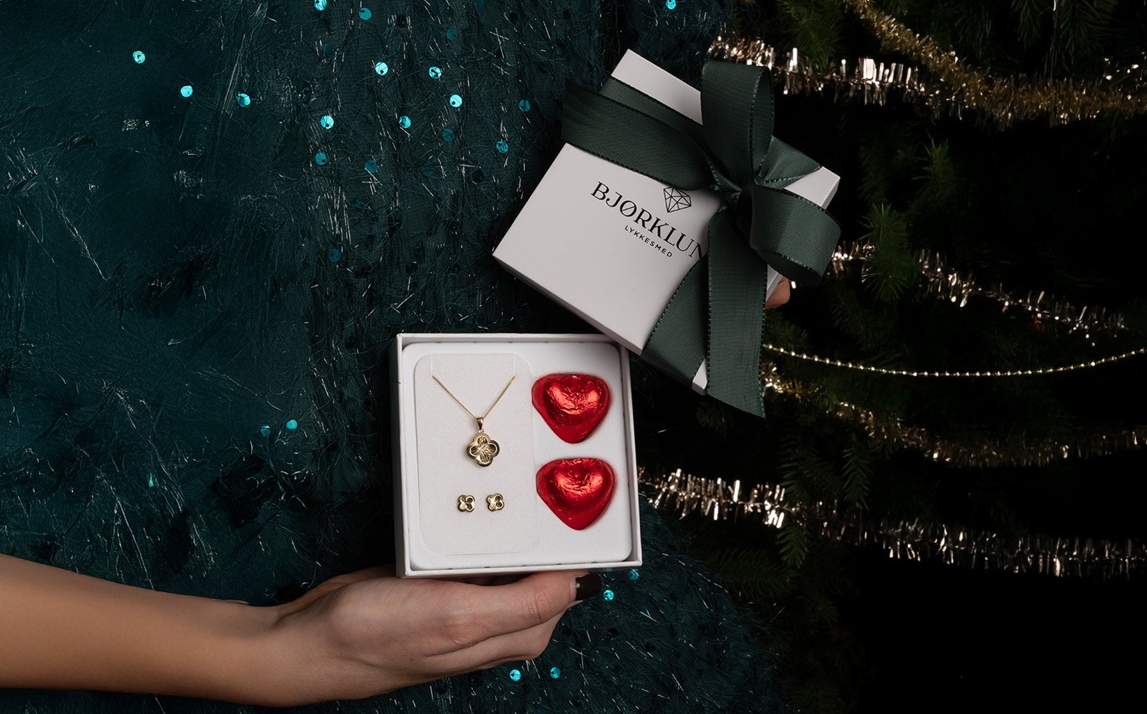 Gavesett til mamma Hender holder Bjørklund-gaveeske med gullsmykkesett og to røde hjerteformede sjokolader foran juletre.