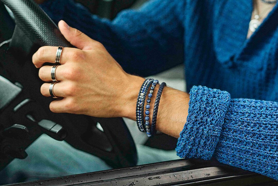 Nærbilde av armen til en mann som holder styret mens hans kjører bil i blå strikka genser og med armbånd og ringer..