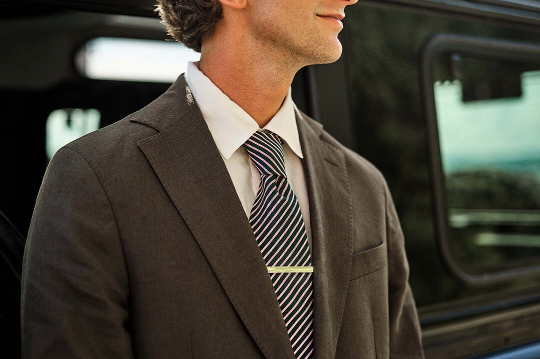 Klokker for menn Mann i dress med stripet slips og gullfarget slipsnål står foran bil med døren åpen i bakgrunnen.