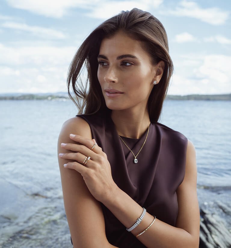 House of Piene diamanter Kvinne med langt hår står ved sjøen iført mørk topp, smykker, ringer, armbånd og øredobber i gull og sølv.
