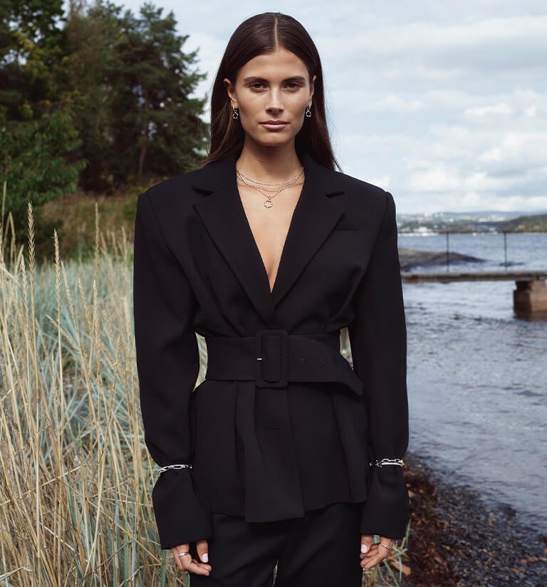 House of Piene - Sølv En ung kvinne med mørkt hår og hvite kape og sølv halskjede, ler og ser til sin høyre, med havutsikt i bakgrunne.