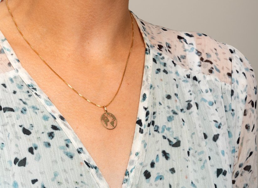 Jordklodesmykker i sølv og gull Nærbilde av en kvinnes hals med et kjede med jordklodesmykke, og hvit bluse med flekker i forskjellige blåfarger.