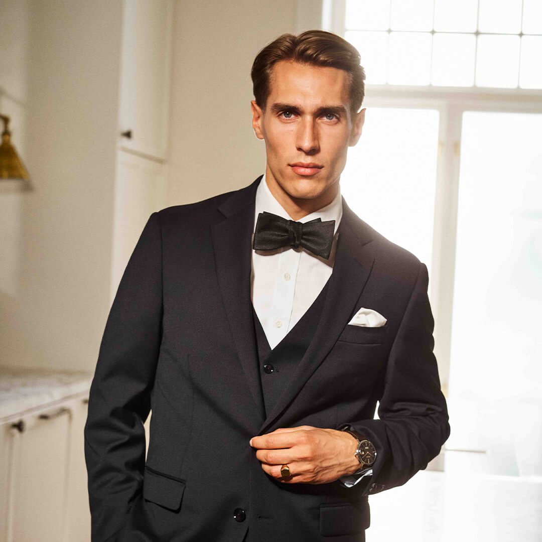 Julegaver til pappa Mann i sort smoking med sløyfe og klokke står ved marmorkamin i lyst rom med store vinduer