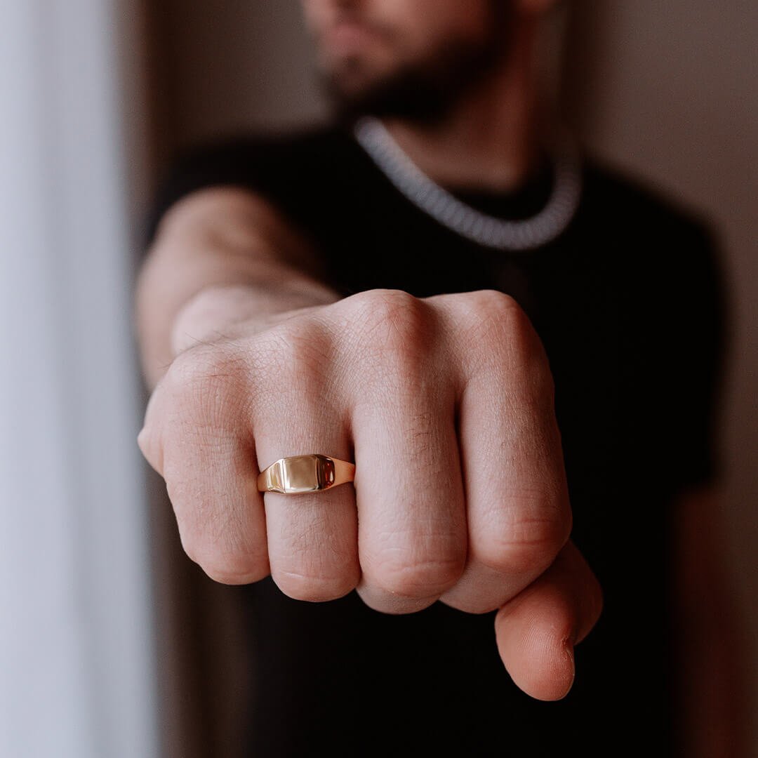 Ringer til menn En mann med skjegg, svart t-shirt og hvitt kjede strekker ut en hånd foran seg med en gullring i fokus, der mannen er i ufokus.