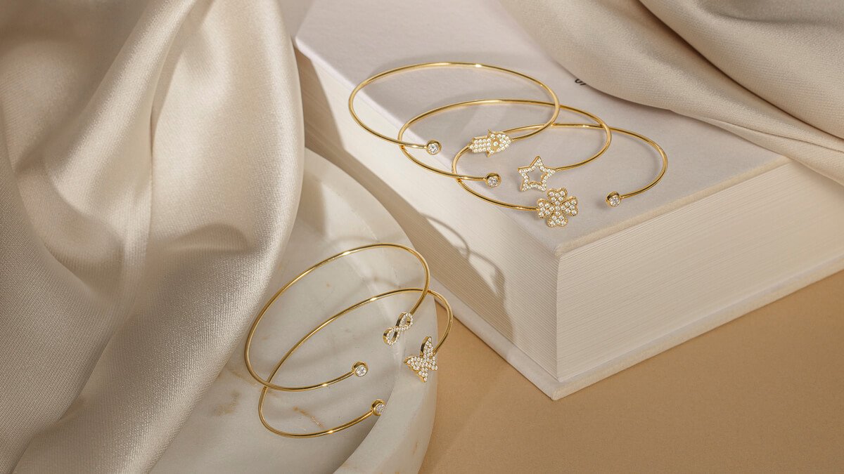 Fem åpne armbånd i gull med ulike symboler og steiner, dandert på bok og beige tekstiler i mykt lys.
