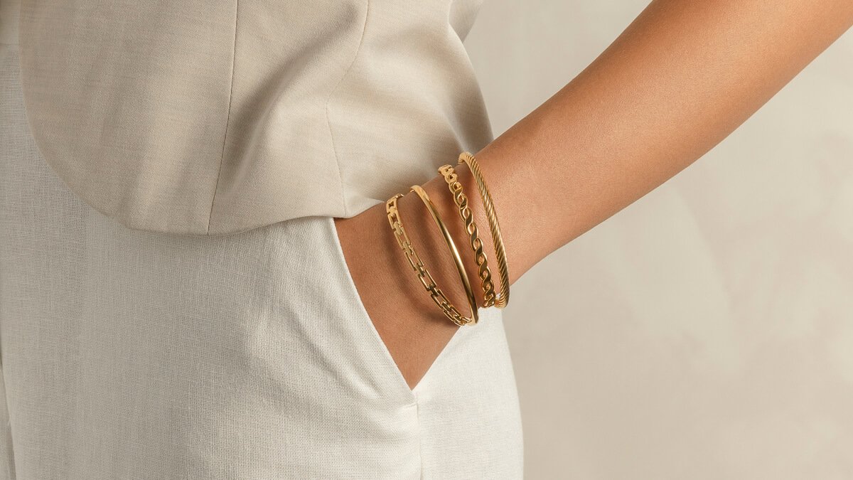 Fire gullfargede armbånd i ulike design på en kvinnearm som hviler i en lomme, med lys beige bakgrunn.