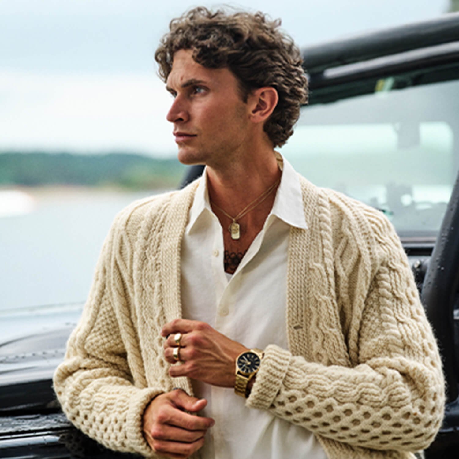 Les Stilguide for menn Mann med krøllete hår iført hvit skjorte og strikket beige cardigan, med klokke, ringer og halskjeder, står ved svart bil med åpent vindu.