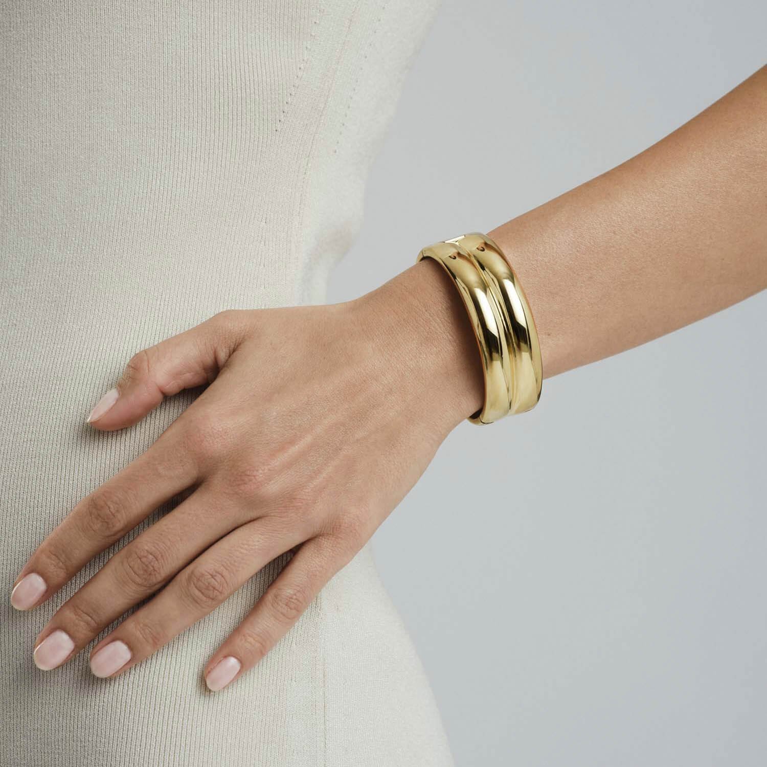 Elegant kvinnearm iført en tykk, gullfarget Mockberg-armbånd, stylet over en lys, tettsittende strikkekjole. Armbåndet har en minimalistisk, bølget design som reflekterer lyset og gir et moderne uttrykk.