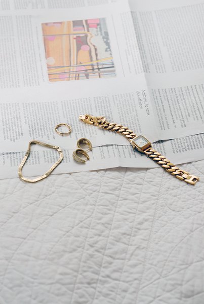 Et armbån, en ring, et par øredobber og en klokke i gull, ligger på et avispapir og et hvitt teppe.
